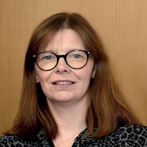 A close-up photo of Dr Ursula Mason standing in front of a wooden background. 